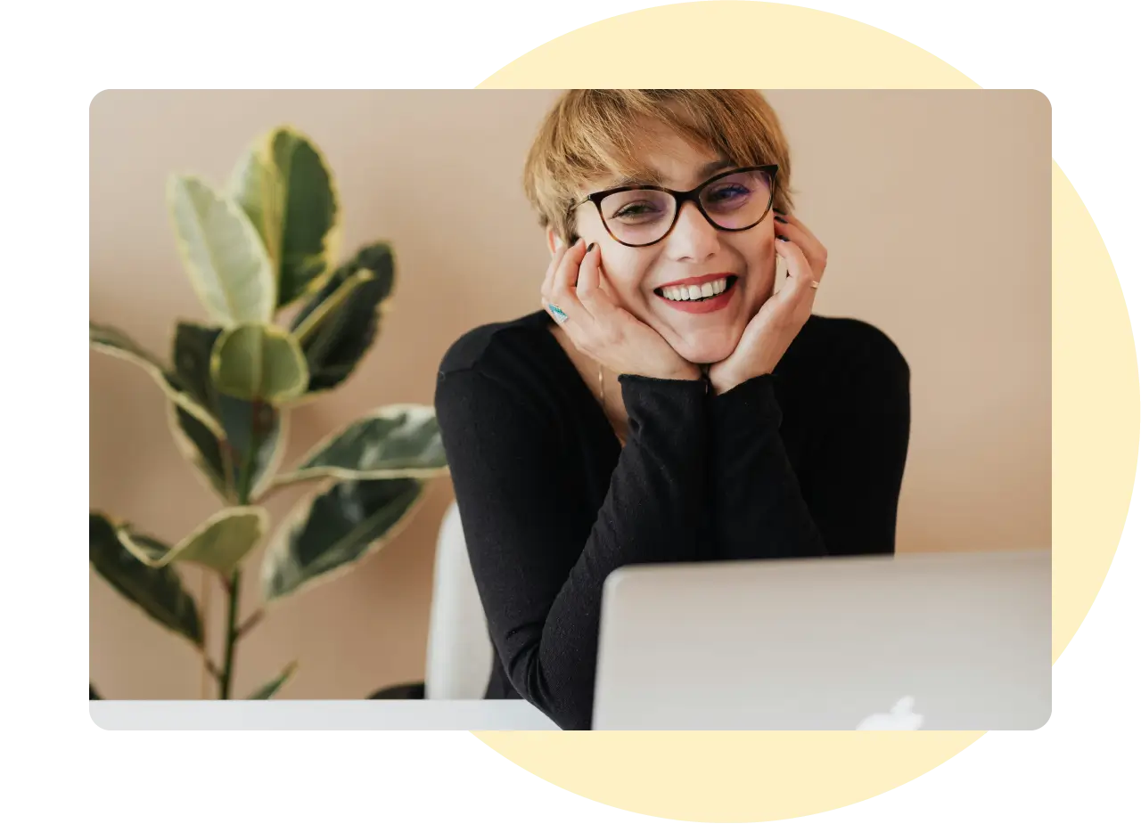 A woman smiling at the camera in front of her laptop. Her head is cupped in her hands and she looks relaxed.