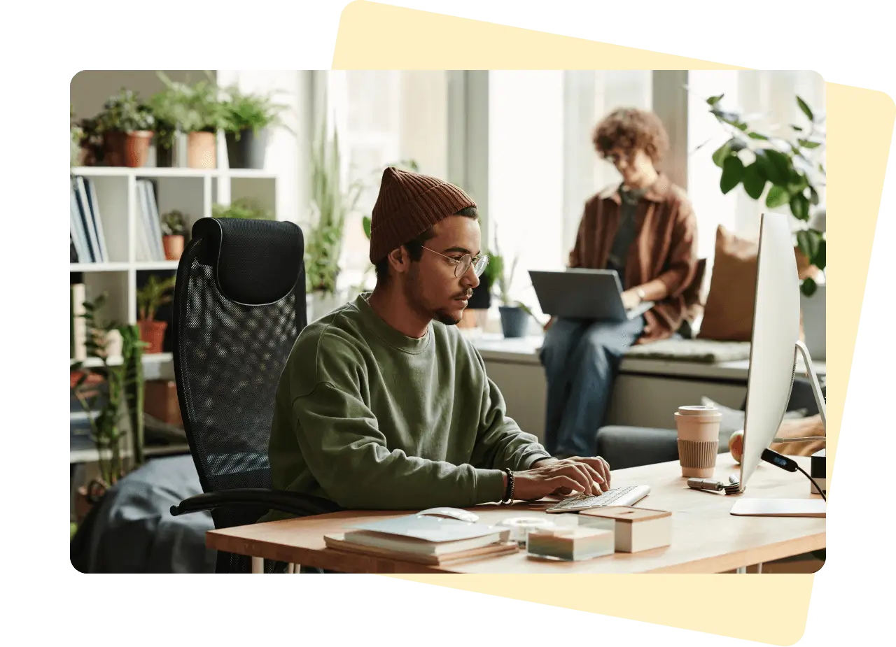 Man wearing a beanie hat is working at a desk on his computer. There is a woman sat on a window sill in the background, using a laptop.