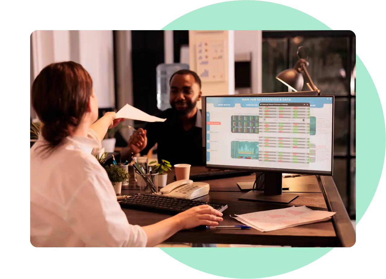 A woman passing a document over her desk to a man sitting on the other side. A analytics dashboard is displayed on her computer screen.