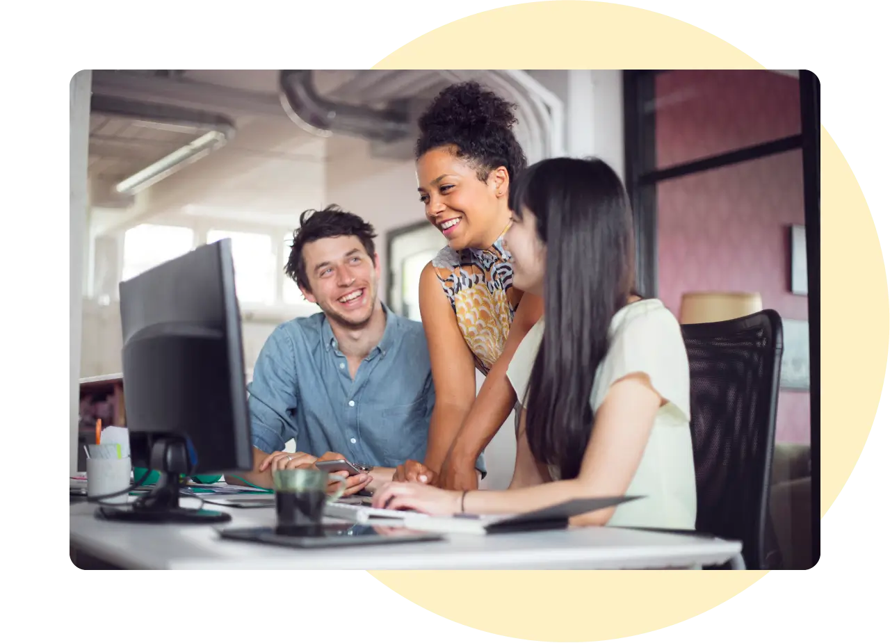 Two women and a man sitting at a desk and smiling at a computer screen in front of them.