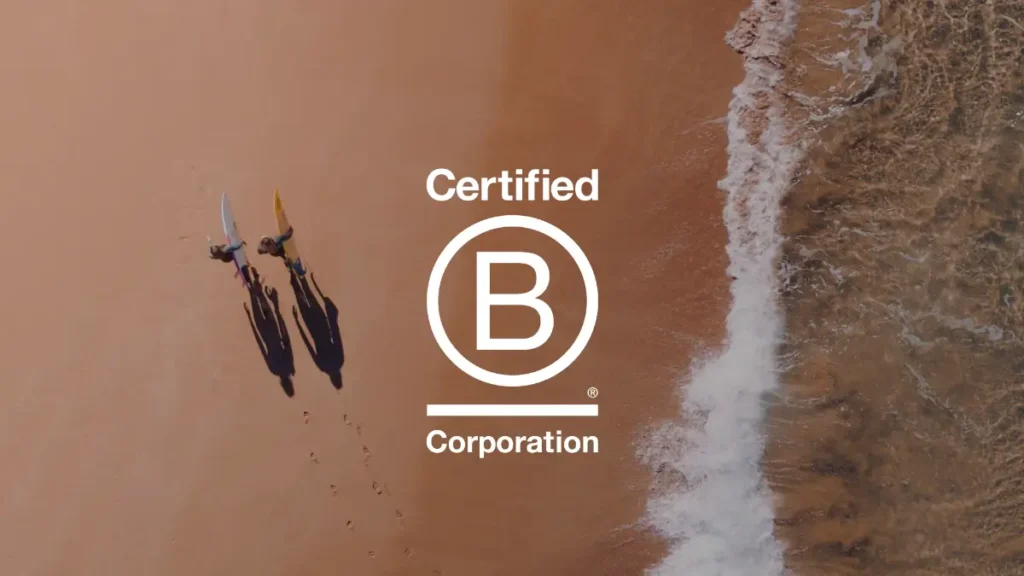 Aerial photo of two people walking along a beach. A wave is breaking on the sand and the Certified B Corp logo is overlaid.