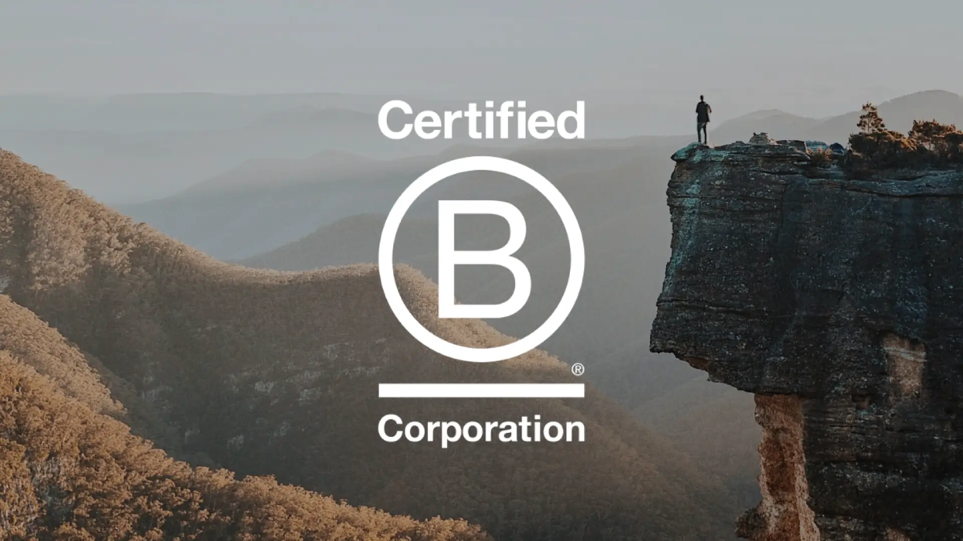 Landscape photo of a person standing on the edge of a cliff, overlooking a valley. On top of the photo is the Certified B Corporate logo.